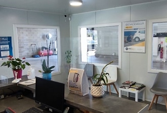 Accueil Garage Auto : Réception Propre et Moderne Accueil d'un garage automobile avec un comptoir, un ordinateur, des plantes, et des baies vitrées donnant sur l'atelier. Un panneau mentionne 'INFO OFFRES SPÉCIALES'.