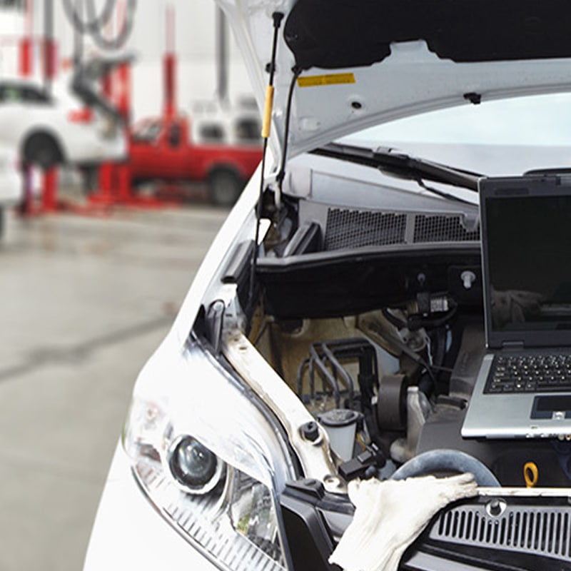 Diagnostic Auto Électronique en Garage : Ordinateur sur Moteur Capot ouvert d'une voiture blanche dans un garage, avec un ordinateur portable posé sur le moteur pour un diagnostic, et des gants de travail à proximité.