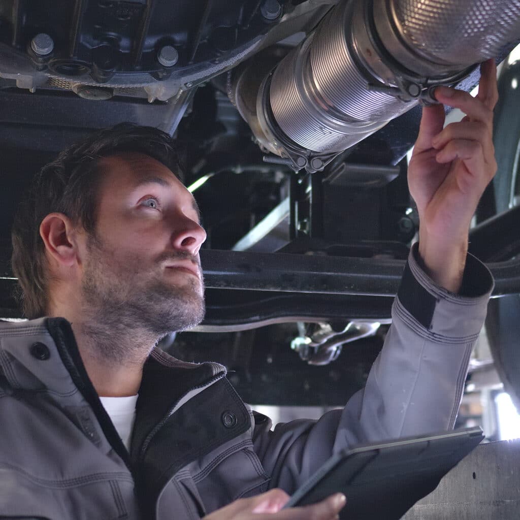 Mécanicien automobile vérifie et ajuste un collier d'échappement Mécanicien vêtu d'une veste grise examinant et ajustant un collier de serrage sur un tuyau d'échappement flexible de voiture avec une tablette à la main.