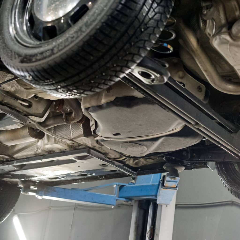 Inspection Sous Voiture : Châssis et Pont Élévateur Vue rapprochée du dessous d'une voiture soulevée sur un pont élévateur bleu dans un garage, montrant le pneu, le châssis et des composants d'échappement.